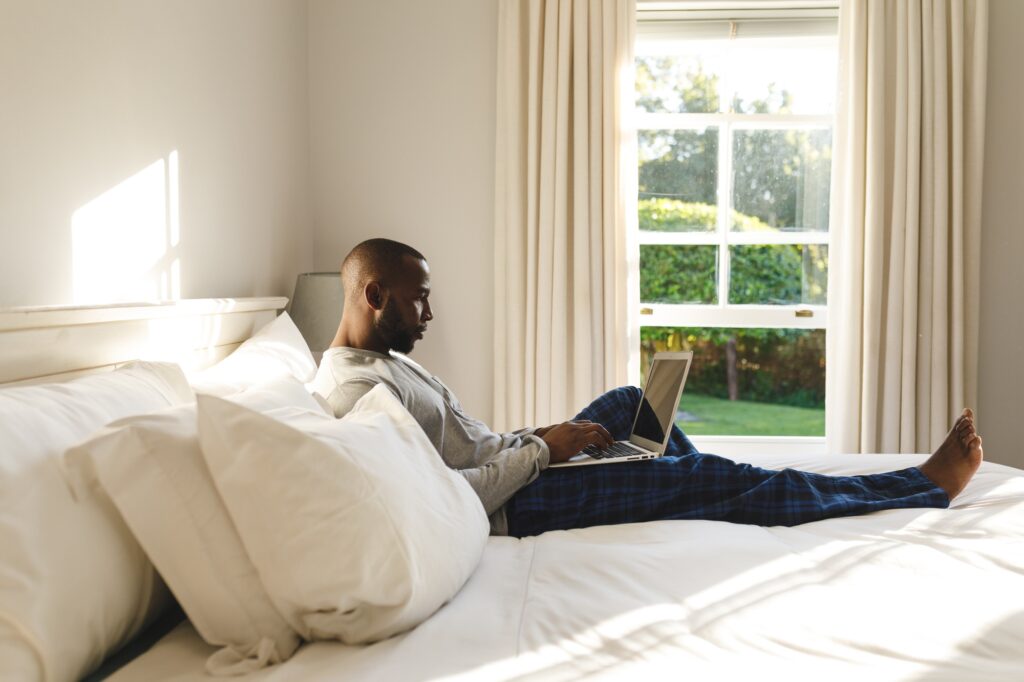 African american man using laptop and lying on bed in his bedroom